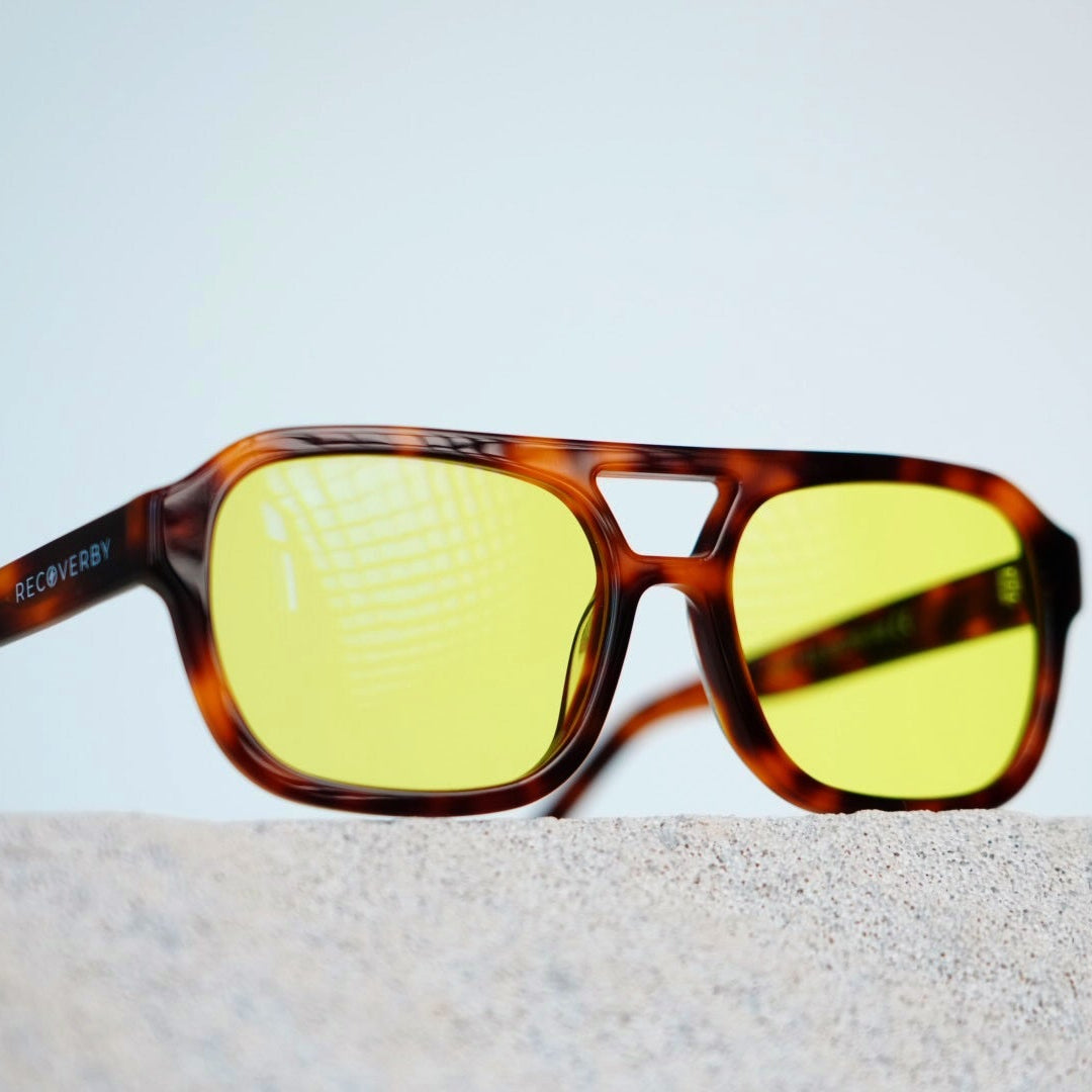 A pair of square sunglasses with a brown tortoiseshell frame and yellow tinted lenses, resting on a light gray textured surface against a pale blue background.