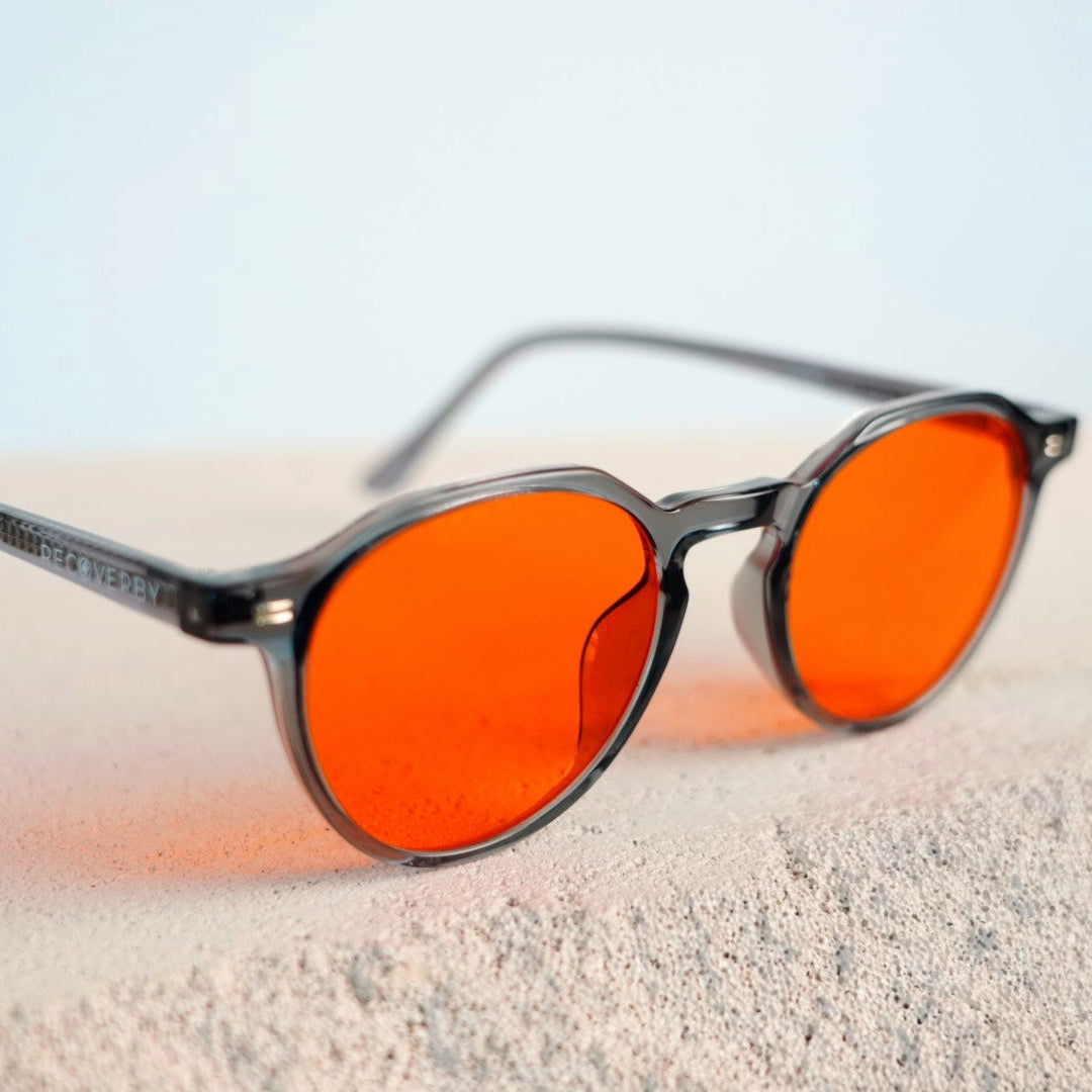 Oversized round sunglasses with vibrant orange lenses and a dark gray translucent frame, resting on a light beige textured surface against a pale blue background.