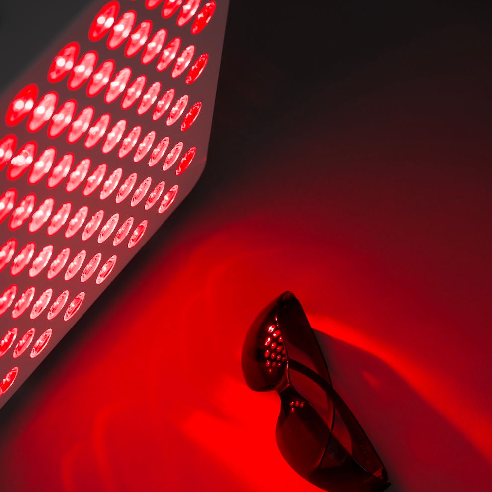 Red light therapy panel emitting a red glow, shown alongside a pair of dark red sunglasses on a dark background.