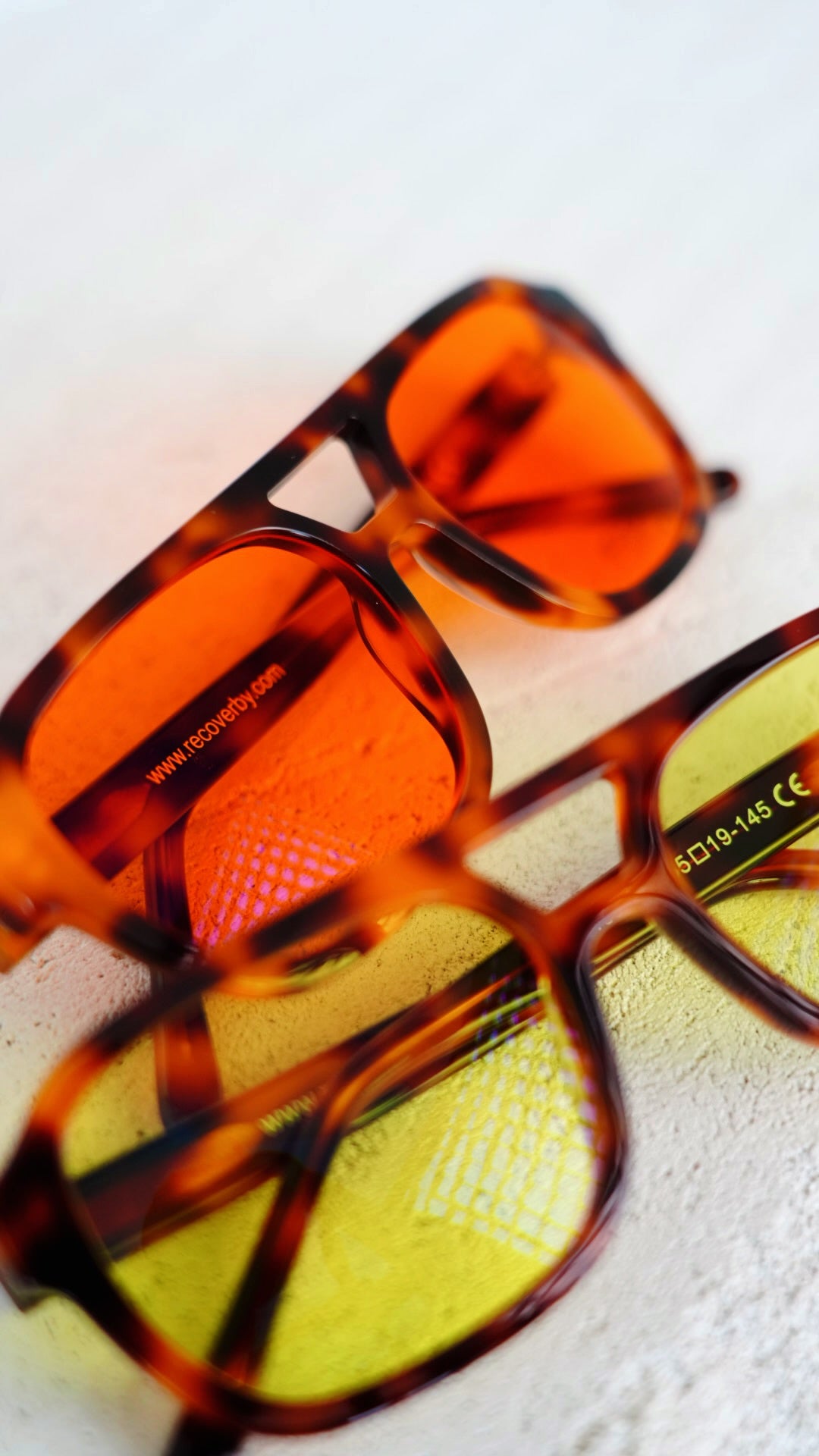 Two pairs of square-frame Blue Light Blocking glasses Recoverby, Day sync and Night Sync Aura frames with tortoiseshell frames, one with orange-tinted lenses and the other with yellow-tinted lenses, resting on a light textured surface.