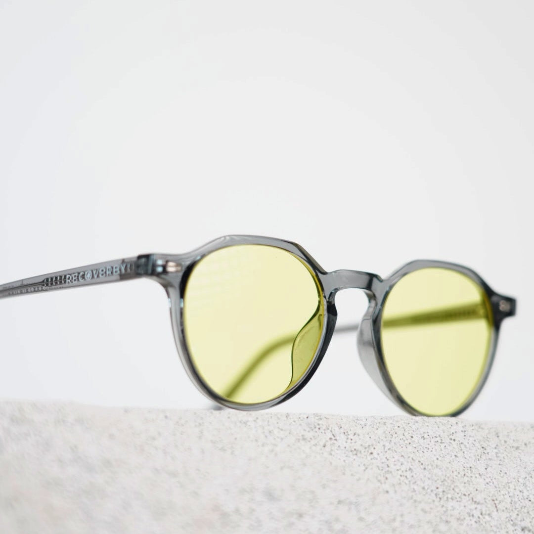 Round blue light blocking glasses with translucent gray frames and yellow tinted lenses, resting on a light textured surface against a white background.