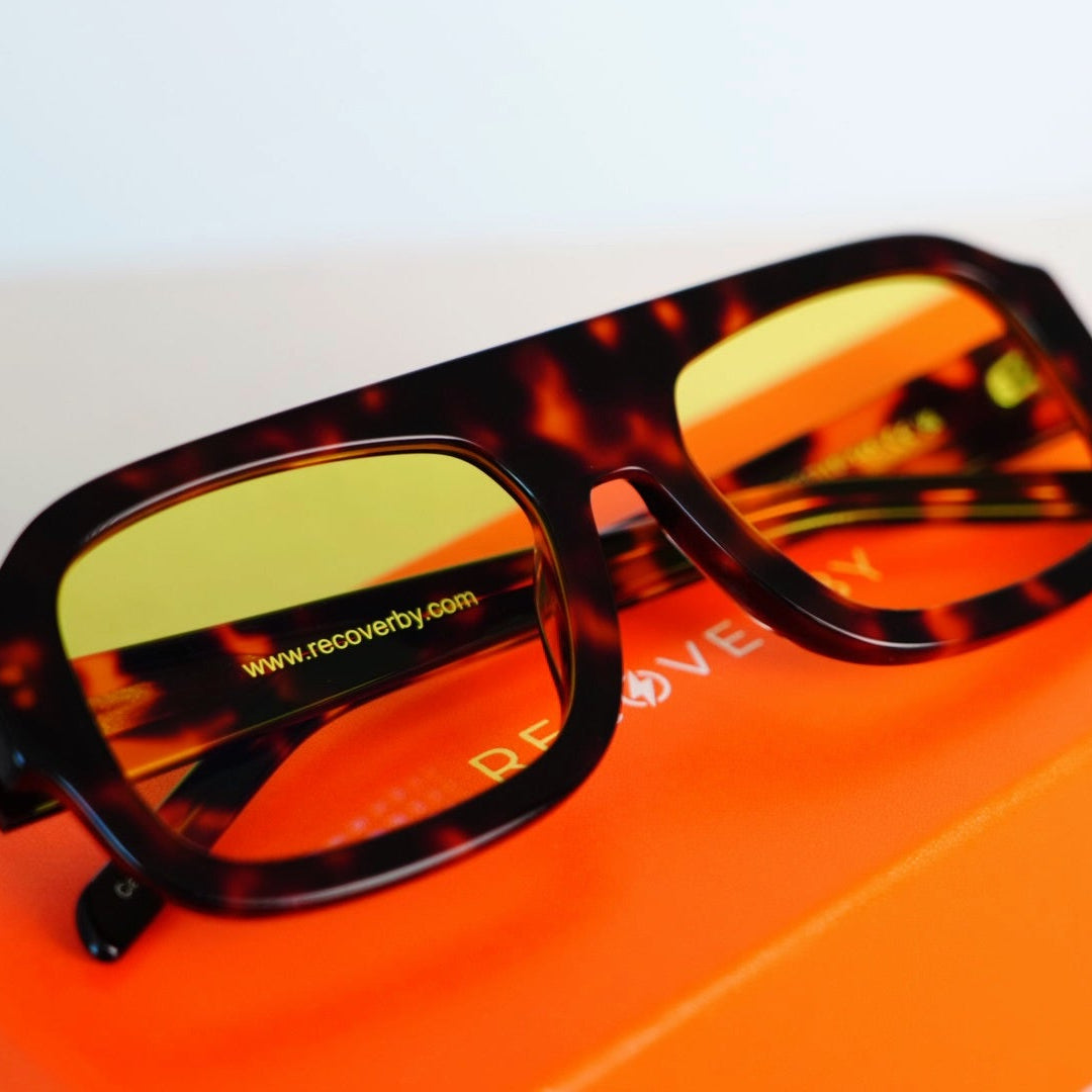 A pair of oversized square sunglasses with a dark tortoiseshell frame and yellow-tinted lenses, resting on a bright orange box against a clean white background.