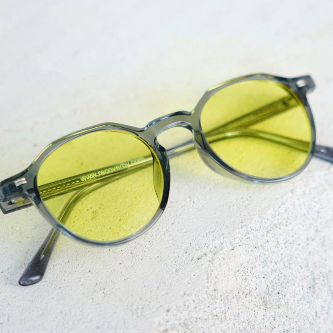 Oversized round sunglasses with translucent smoky gray frames and yellow tinted lenses, resting on a light textured surface.