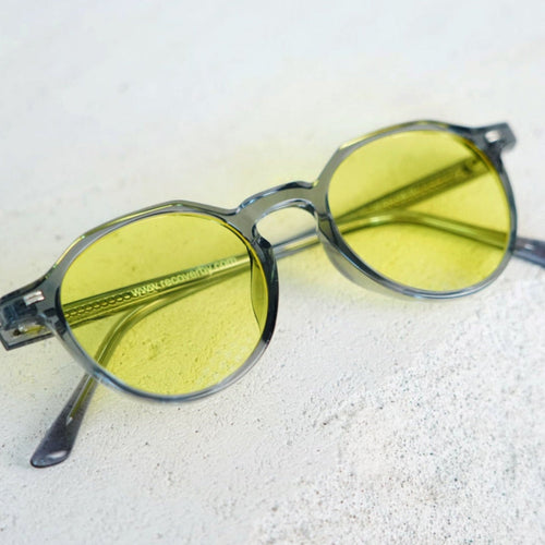 Oversized round sunglasses with translucent smoky gray frames and yellow tinted lenses, resting on a light textured surface.