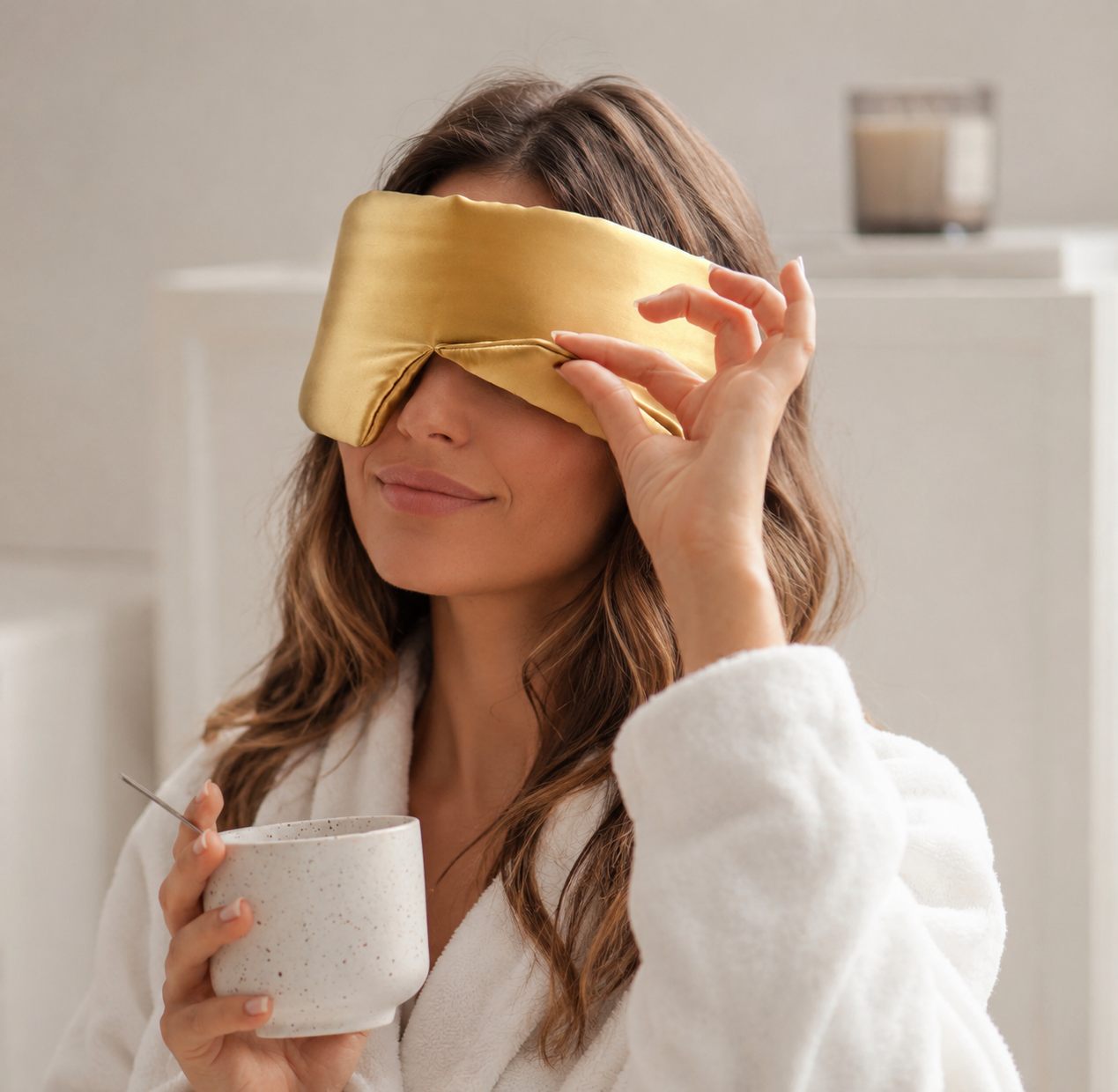 A woman in a white robe wearing a satin gold sleep mask and holding a speckled white mug in a bright, modern bathroom setting.
