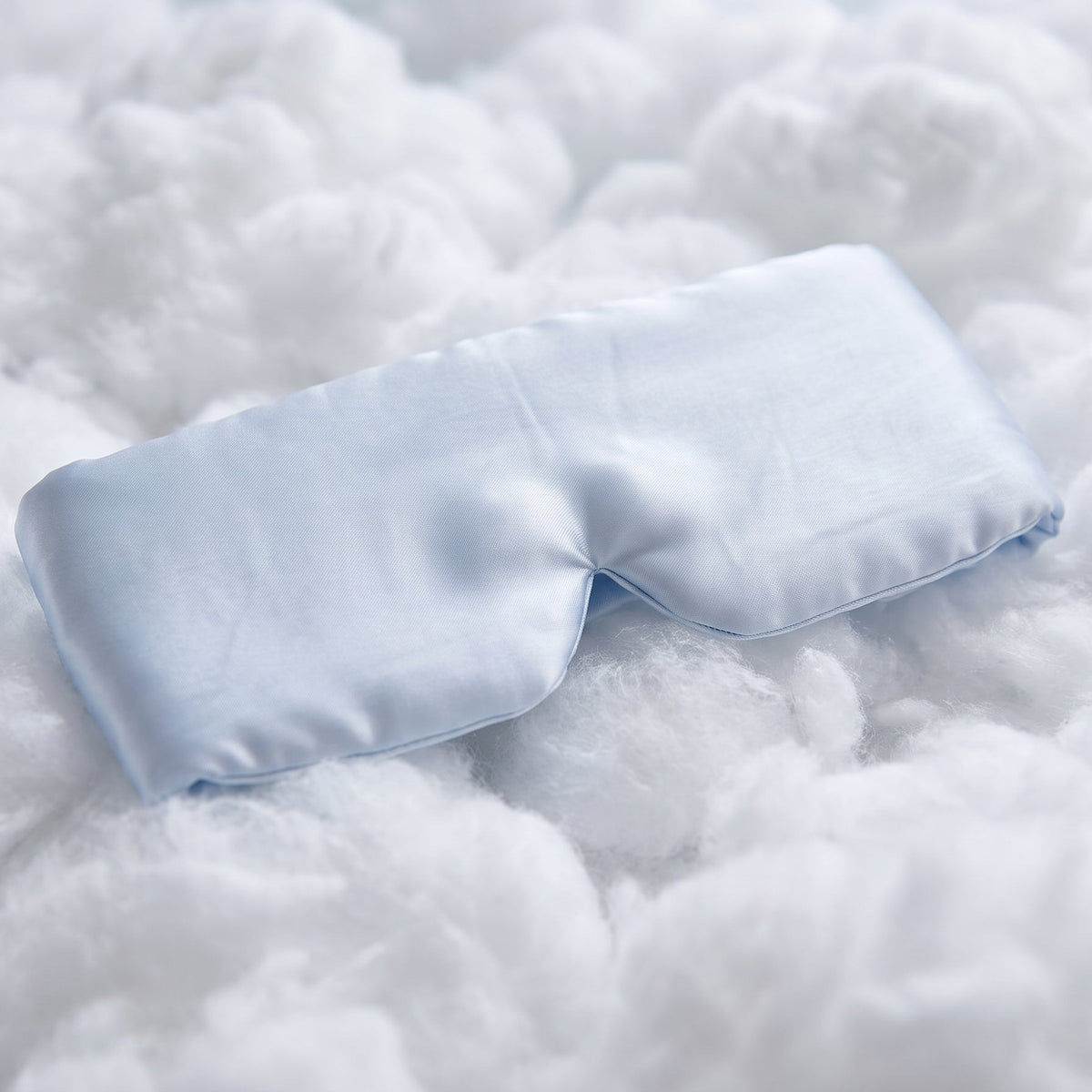 A light blue satin sleep mask resting on a soft, fluffy white textured blanket.