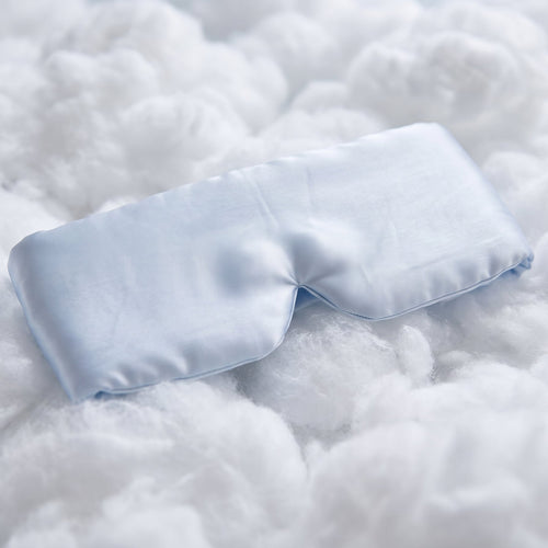 A light blue satin sleep mask resting on a soft, fluffy white textured blanket.