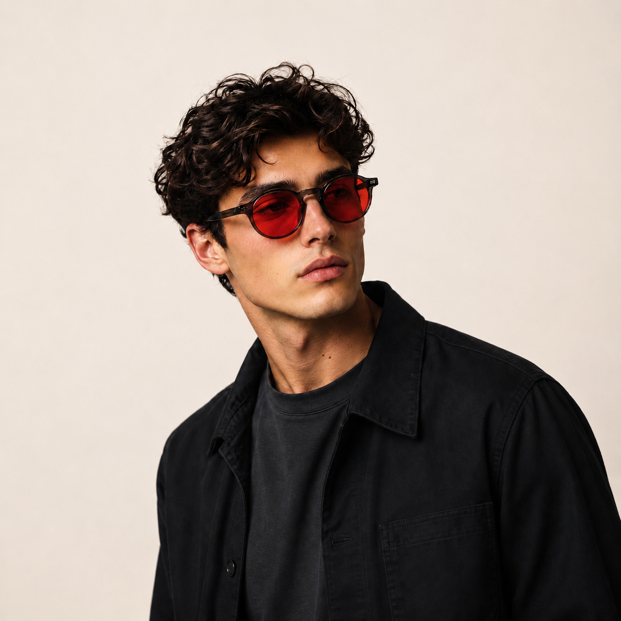A man with curly dark hair wearing tortoiseshell RECOVERBY BLUE LIGHT BLOCKING GLASSES with red lenses, styled with a black shirt over a dark gray t-shirt, against a plain beige background.