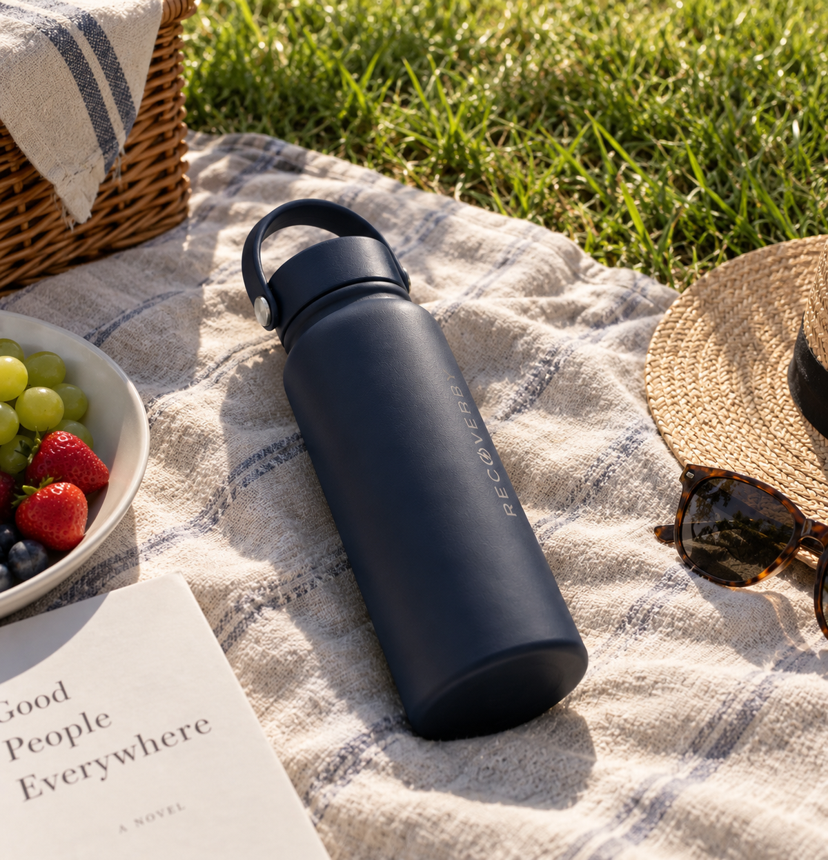 A matte navy blue water bottle with a carry loop and subtle vertical branding, placed on a white plaid picnic blanket outdoors on grass. A wicker basket, bowl of fruit, sunglasses, and a book are visible around it.