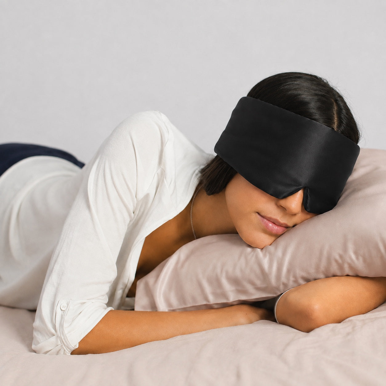 A woman lying on a bed with her eyes closed, wearing a black satin sleep mask and a white long-sleeve shirt.
