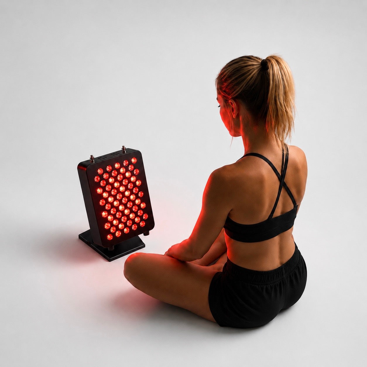 A woman in a black sports bra and shorts sits cross-legged on a light gray studio floor, facing a black Red light Led therapy panel from recoverby.com that glows with red light.
