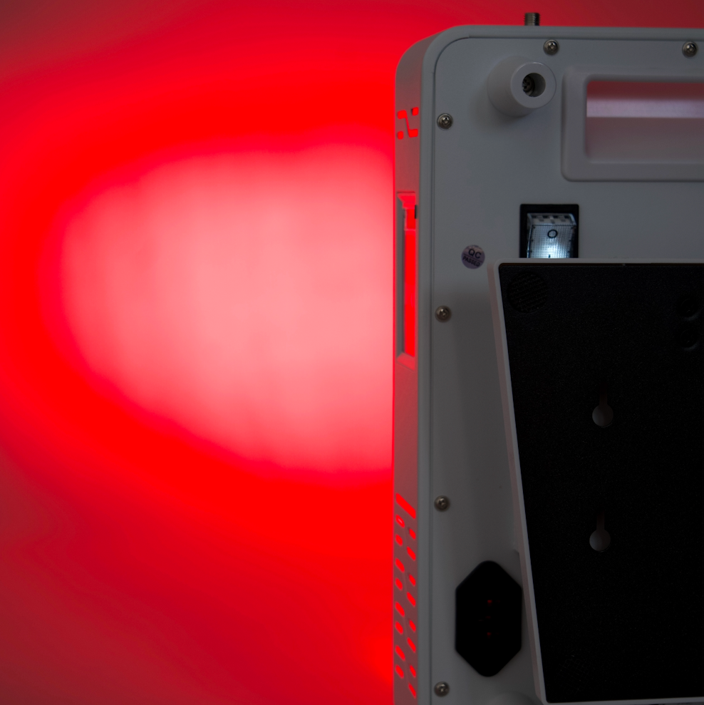 A white electronic device shown from the back, emitting a strong red light that illuminates the background. The device has a white plastic casing with a black panel on the back, screws, and a small illuminated component.