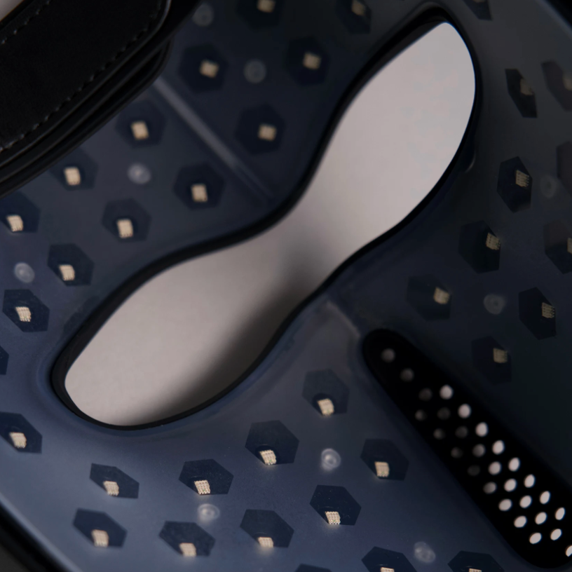 Close-up of a red light face mask inside led lights, a honeycomb pattern of small metallic gold dots and cutouts, set against a neutral light gray background.