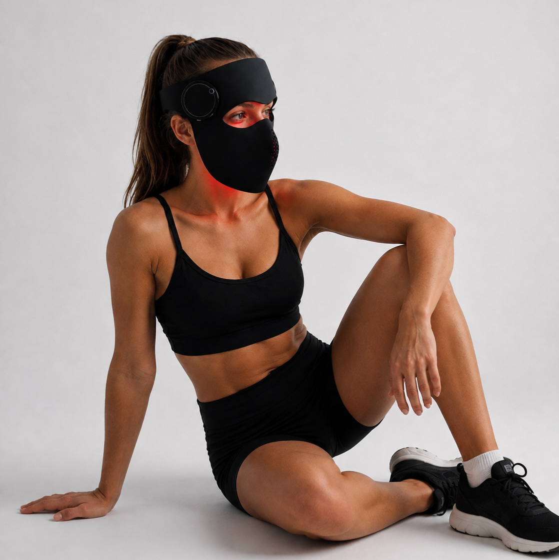 A woman in a Recoverby red Light mask GO and in black sports bra and shorts sits on a light gray studio floor wearing a black athletic face mask with a built-in headband and small circular visor, against a plain light gray background.