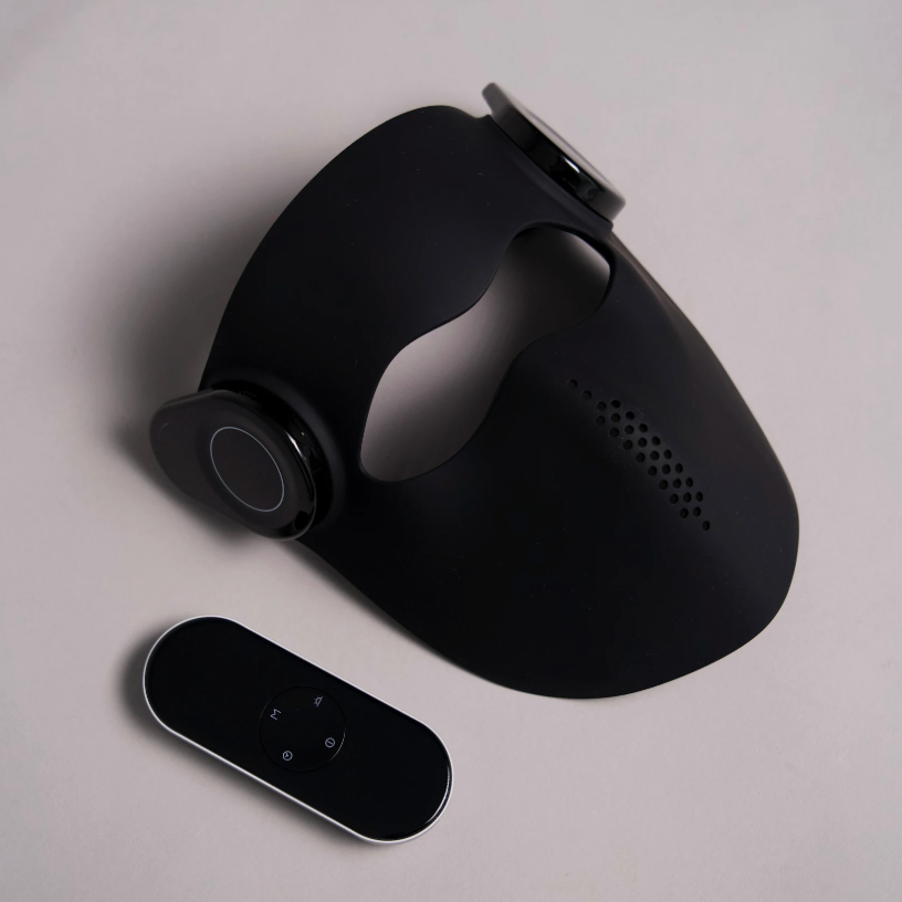 A matte black wearable Red light Face mask device with a curved ergonomic shape and a small oval remote control, photographed on a clean light gray background.