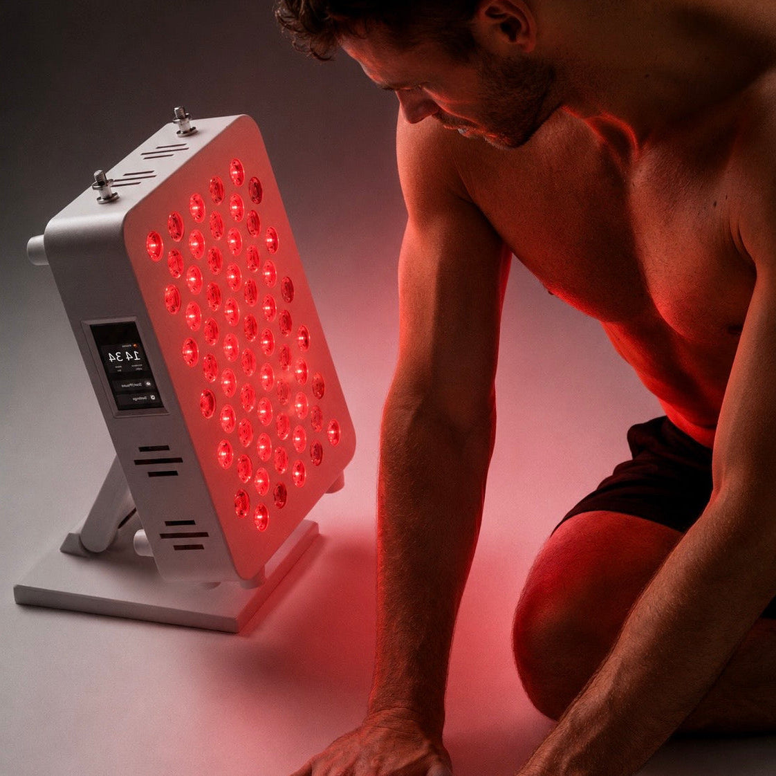A man kneels beside a white LED light therapy panel that is glowing with red light, set against a dark studio background.