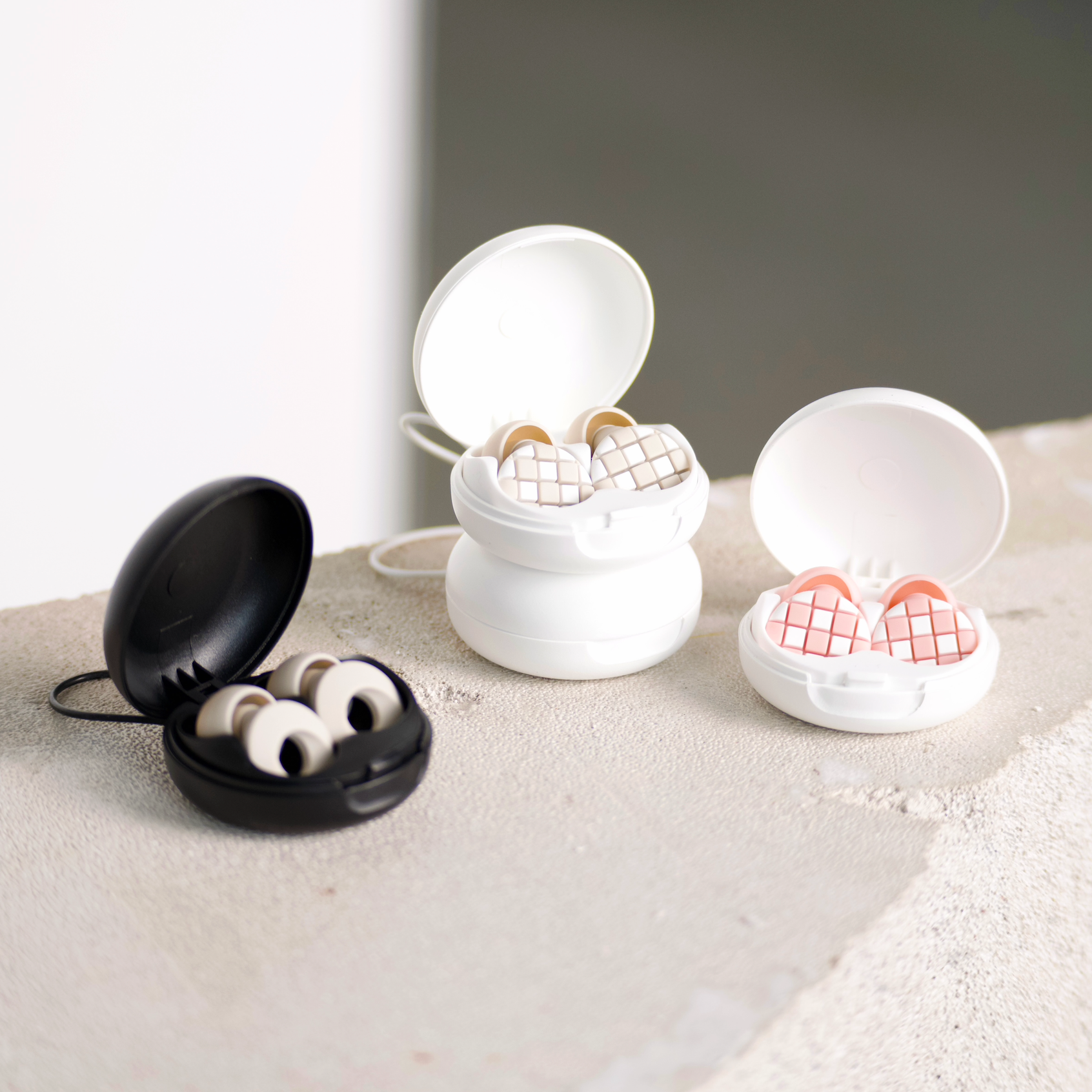 Three pairs of in-ear earplugs in open round cases, shown on a textured light beige surface against a soft gray background. One case is black, and the other two are white; the earplugs inside are beige with a grid pattern and one pair has pink patterned earplugs.