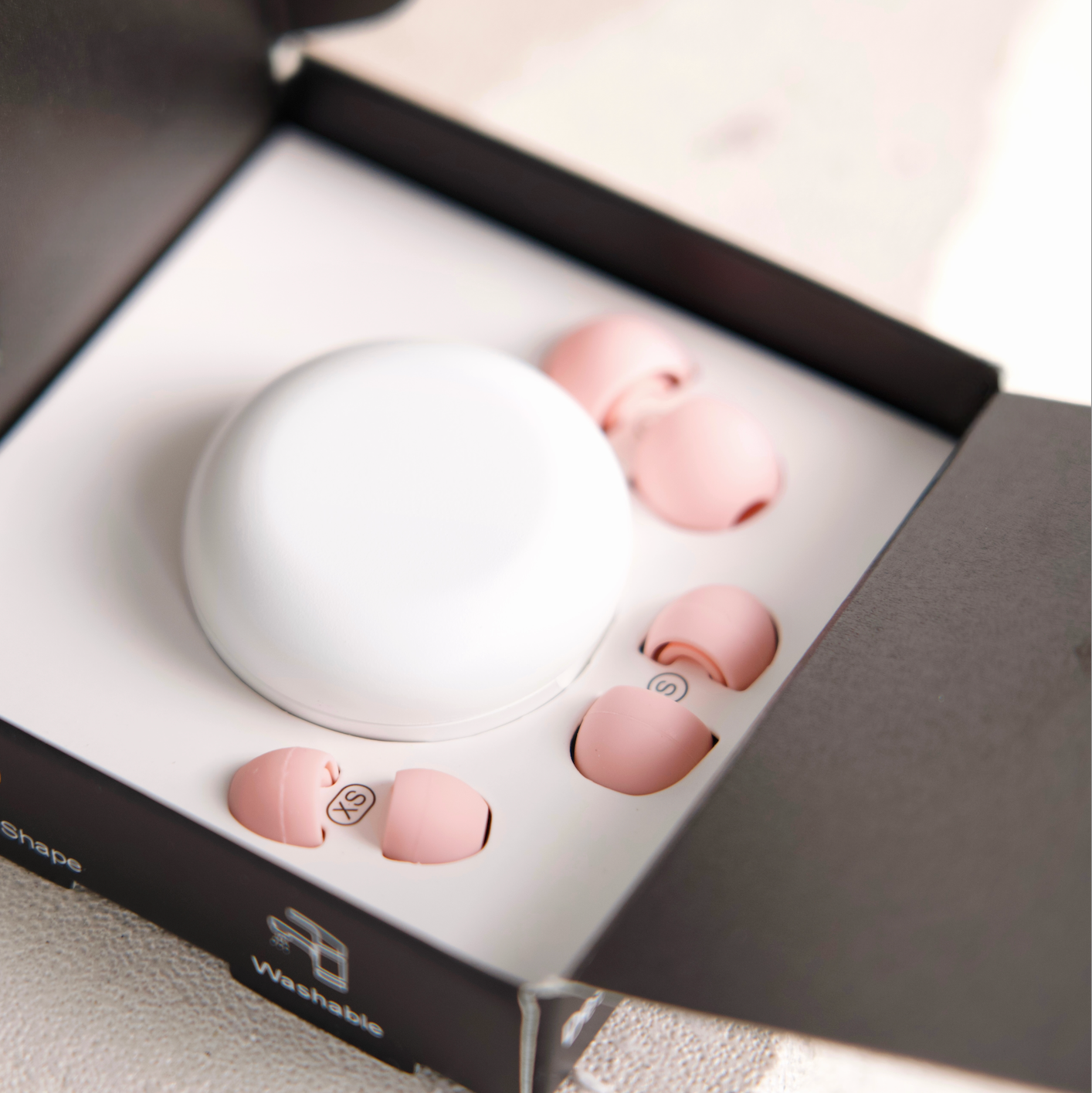 A pair of small pink earbuds arranged around a white oval charging case inside an open black presentation box on a light surface.