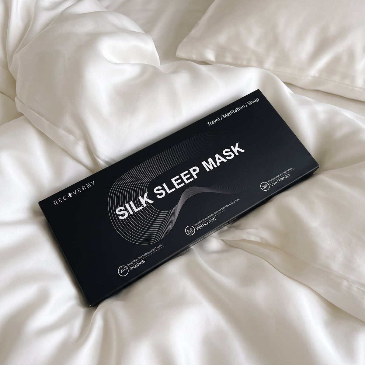 A black rectangular box labeled 'SILK SLEEP MASK' lies on rumpled white bedding with pillows visible in the background from recoverby 