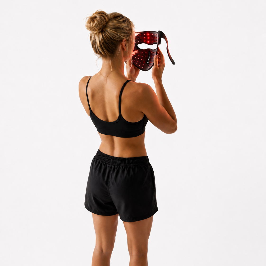 A woman viewed from behind wearing a black sports bra and black athletic shorts, holding a red and black patterned face mask up to her face against a plain white studio background.